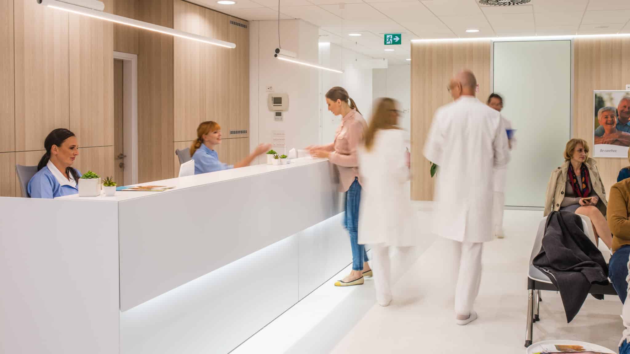 Blurred motion of doctors walking in a busy hospital reception area, patients sitting on chair.