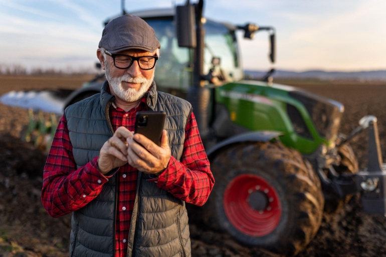 Farmer using mobile UCaaS solutions on a smartphone in front of a tractor in a field